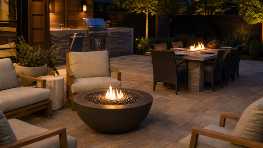 A low rectangular fire table with a gentle linear flame sits between a sectional seating area and a stone patio, with an outdoor kitchen in the background.